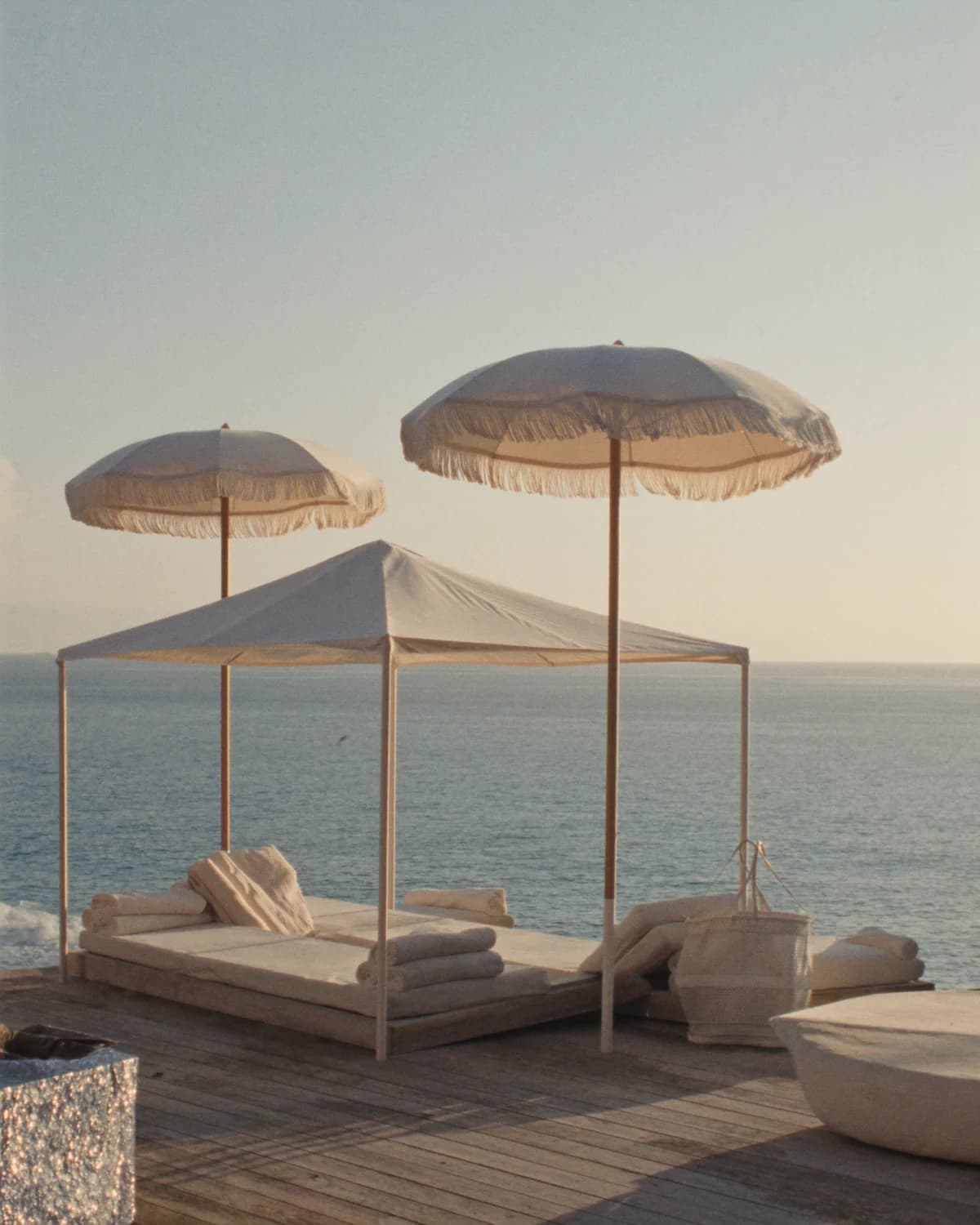 An ivory canopy, daybeds and fringed shade umbrellas arranged for a calm coastal morning