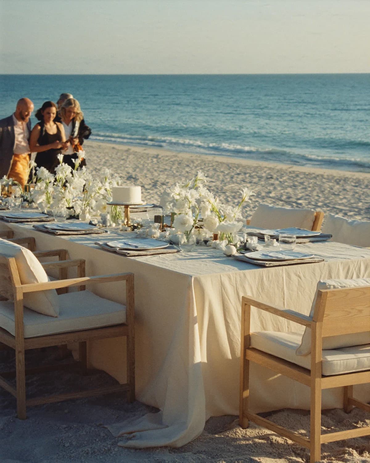 A long linen celebration table with white flowers, pale wood chairs and guests beside the ocean